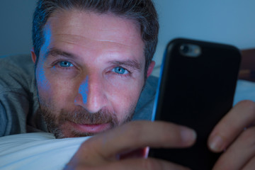 Wall Mural - happy man with blue eyes lying on bed late at night in dark light networking on mobile phone or online dating smiling relaxed enjoying internet in social media addiction