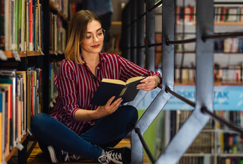 Wall Mural - Young female student study in the library reading book while sitting near bookshelf.