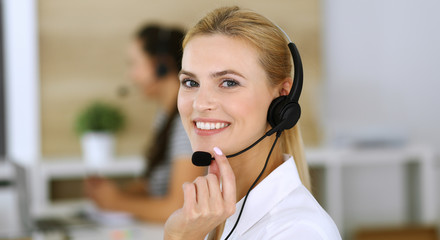 Blonde business woman using headset for communication and consulting people at customer service office. Call center. Group of operators at work at the background.