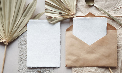 Wall Mural - Blank white Wedding invitation cards mockups with dried palm leaf and craft envelope on textured table backgound. Elegant modern template for branding identity. Tropical design. Flat lay, top view