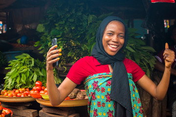 Wall Mural - African woman selling food stuff in a cheerful local market feeling excited and happy expressing raising both hands doing a thumbs up, also holding her mobile phone, smiling