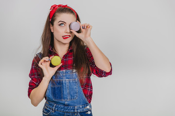 Beautiful happy cute young pretty woman holding two macaroons, hiding her eye behind blue one, twisting her lip.