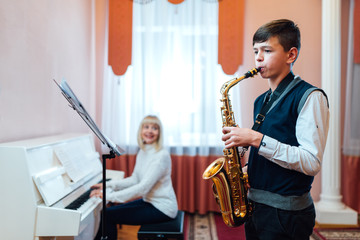 A teenage boy learns to play the saxophone in a music lesson to the accompaniment of a female teacher on the piano.