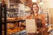 Happy mature female baker smiling, holding out paper shopping bag to the camera, selling delicious bread and pastry