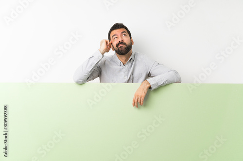 Fotografía Young handsome man with beard holding a big green empty placard having doubts an
