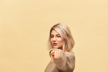 Cute charming young blonde woman in cashmere sweater reaching out hand and pointing index finer at camera, choosing you, inviting to dance with her, having energetic enthusiastic look, smiling
