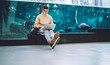 © GalakticDreamer - Young man using laptop and sitting with legs crossed near aquarium