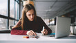 © GalakticDreamer - Young attractive female writer scribing notes to textbook prepare for exam, girl doing homework sitting desktop with modern laptop computer, woman making plan for tomorrow and creating  to do list