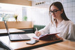 © GalakticDreamer - Positive female student in eyeglasses enjoying time at university campus while noting funny things of day, young hipster girl writing impression review of visiting cafeteria in textbook indoors
