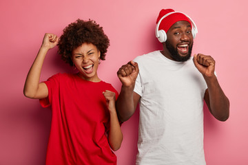 Sticker - Happy woman and man dance together, listen music in stereo headphones, keeps hands raised, dressed in casual wear, dance against pink studio background, have friendly relationship, sing along