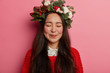 Gentle charming woman with long dark hair poses with eyes closed, smiles pleasantly, imagines something nice, has long straight hair, wears spruce holiday wreath around head, stands over rosy wall