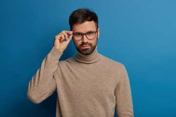 Wall Mural - Handsome self confident man with thick bristle, keeps hand on rim of glasses, wears casual brown turtleneck, looks seriously at camera, has conversation with colleague, discuss their future plans