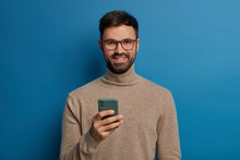 Happy Bearded Male Blogger Rejoices Having New Followers In Blog, Gets Message On Mobile Phone, Has Friendly Smile, Wears Turtleneck, Isolated On Blue Wall, Creats New Post, Uses New Application