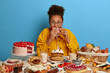 Funny glutton woman bites cakes with big appetite, cant stop eating sweet desserts, being in mood for enjoying sugary freshly baked bakery, surrounded by sweet treasures, has yummy snack, feels hunger