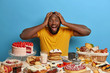 © WHstudio Leushin N - Emotive dark skinned bearded guy keeps both hands on head, opens mouth from surprisement as sees too much desserts at one table, shocked his wife baked lots of confectionery for festive event