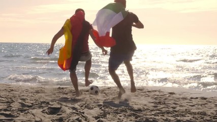 Wall Mural - Two young men wearing italian and german flags as capes play football at the beach on the sea shore at sunset steadycam gimbal dolly