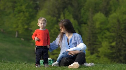 Wall Mural - Mother sit on grass, hold newborn baby and caress blond hair child, happy in nature