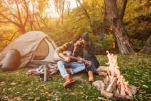 Young Loving Couple Of Tourists Relaxing Near The Bonfire In The Nature..Handsome Man Playing Guitar Near Tent. Beautiful Woman Listening To Music On A Hike By The Campfire.