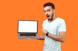 Portrait of excited shocked surprised brunette man with beard in white t-shirt pointing at laptop with blank screen, empty place for internet advertising. studio shot isolated on orange background