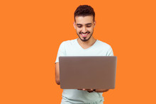 Portrait Of Cheerful Young Brunette Man With Beard In White T-shirt Typing Email On Laptop And Smiling, Reading Positive Message Or Surfing The Web. Indoor Studio Shot Isolated On Orange Background
