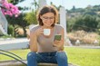 Mature woman resting outdoor reading smartphone with cup