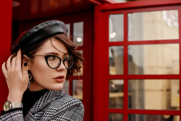 Fashionable, elegant woman wearing glasses, trendy leather beret, hand watch, classic autumn coat, posing near red call box in street of city. Copy, empty space for text