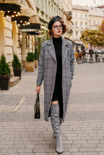 Outdoor Full-length Fashion Portrait Of  Elegant Woman Wearing Classic  Checkered Midi Coat, Short Turtleneck Dress, Gray Suede High, Over Knee Boots, Beret, Glasses,walking In Street Of European City