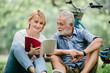 © May Chanikran - Happy elderly couple with smiling face enjoying together, reading a book in the park, spending time and relaxing time concept.