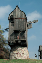 A Woman Next To A Large Abandoned Windmill. A Female Photographer With A Backpack Stands Next To A Huge Wooden Mill