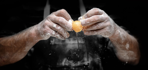 Baker chef breaks yellow egg into fresh dough on kitchen table, black background