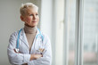 © Seventyfour - Waist up portrait of mature female doctor standing with arms crossed by window in clinic and looking away pensively, copy space