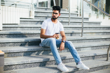 Wall Mural - Young handsome indian man drink coffee while sitting on the stairs in the street