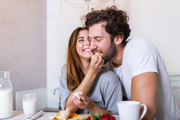 Beautiful young couple is feeding each other and smiling while cooking in kitchen at home. Happy sporty couple is preparing healthy food on light kitchen. Healthy food concept.