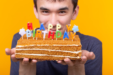 Wall Mural - Close-up of a funny positive guy with a paper cap and glasses holding a congratulatory homemade cake in his hands on a yellow background. Concept and fun and celebration.