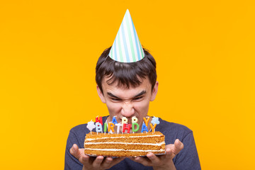 Wall Mural - Portrait of a funny positive guy with a paper cap and glasses holding a congratulatory homemade cake in his hands on a yellow background. Concept and fun and celebration.