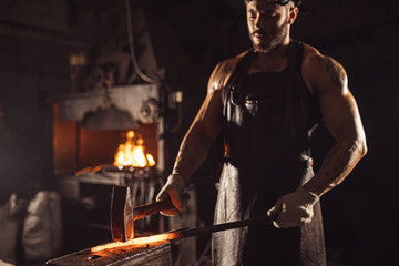 Wall Mural - young caucasian forger beating hot metal using hammer isolated in dark space workshop, performing blacksmith work