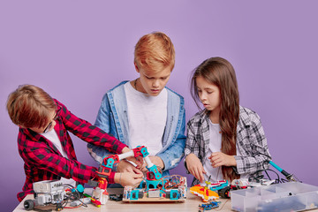 portrait of enthusiastic three kids working as team, assembling robots. three future engineers look at table full of robotics, two boys and one girl