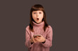 Shocked by an unexpected surprise, a girl on a gray background is holding a mobile phone in her hands and saw something on the Internet. Secure connection and protection from advertising for children