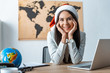 © LIGHTFIELD STUDIOS - smiling travel agent in santa hat looking at camera while sitting at workplace