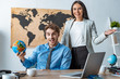 © LIGHTFIELD STUDIOS - attractive travel agent showing welcome gesture while standing near smiling colleague holding globe