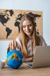 © LIGHTFIELD STUDIOS - smiling travel agent touching globe while sitting at workplace