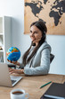 © LIGHTFIELD STUDIOS - smiling travel agent holding globe while sitting at workplace