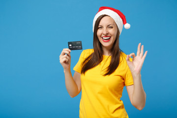 Wall Mural - Cute young woman Santa girl in yellow t-shirt Christmas hat posing isolated on blue background. New Year 2020 celebration holiday concept. Mock up copy space. Hold credit bank card showing OK gesture.