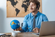 © LIGHTFIELD STUDIOS - smiling travel agent holding globe while sitting at workplace