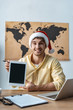 © LIGHTFIELD STUDIOS - cheerful travel agent in santa hat looking at camera while holding digital tablet with blank screen
