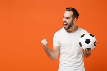 Canvas Print - Joyful young man in casual white t-shirt posing isolated on orange background studio portrait. People sincere emotions lifestyle concept. Mock up copy space. Holding soccer ball doing winner gesture.