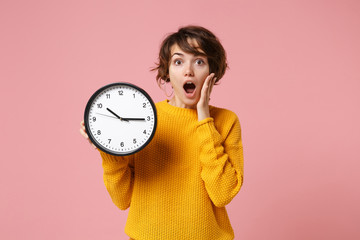 Shocked young brunette woman girl in yellow sweater posing isolated on pastel pink wall background. People sincere emotions lifestyle concept. Mock up copy space. Holding clock, putting hand on cheek.