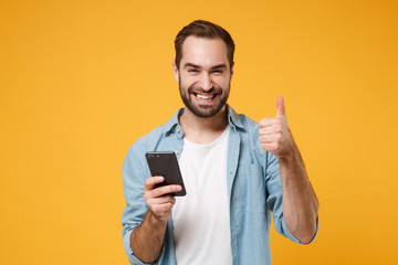 Wall Mural - Smiling young man in casual blue shirt posing isolated on yellow orange wall background in studio. People lifestyle concept. Mock up copy space. Using mobile phone typing sms message showing thumb up.
