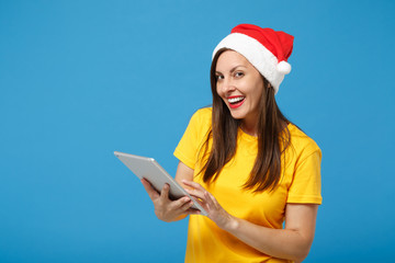 Wall Mural - Cheerful young brunette woman Santa girl in yellow t-shirt Christmas hat posing isolated on blue background. New Year 2020 celebration holiday concept. Mock up copy space. Holding tablet pc computer.