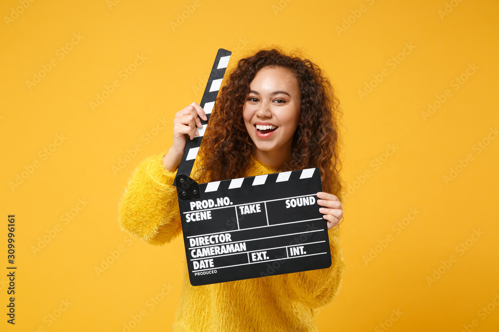 Cheerful young african american girl in fur sweater posing isolated on yellow orange background in studio. People lifestyle concept. Mock up copy space. Holding classic black film making clapperboard. - obrazy, fototapety, plakaty 